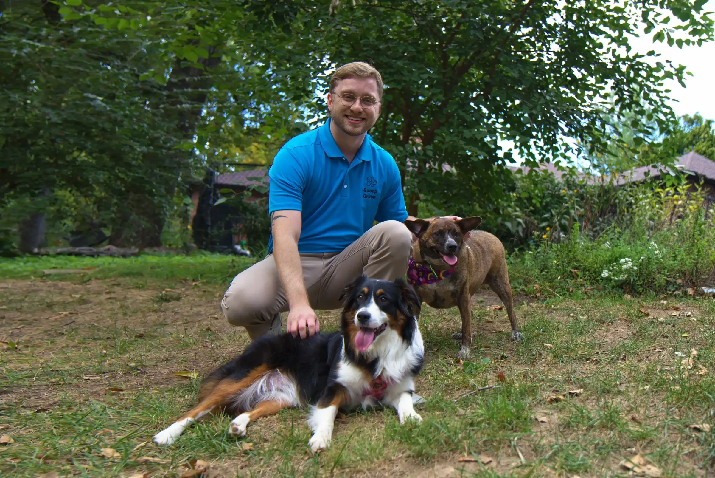 Scoop Group owner Zane Creek in a branded blue polo kneeling in a Richmond Heights, MO backyard with a tricolor Australian Shepherd and a brindle mixed-breed dog